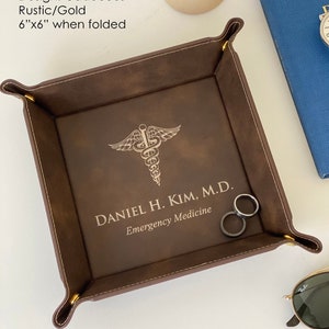 May include: A brown leather valet tray with gold stitching and a gold metal corner piece. The tray features a gold embossed caduceus symbol and the text "DANIEL H. KIM, M.D. Emergency Medicine".
