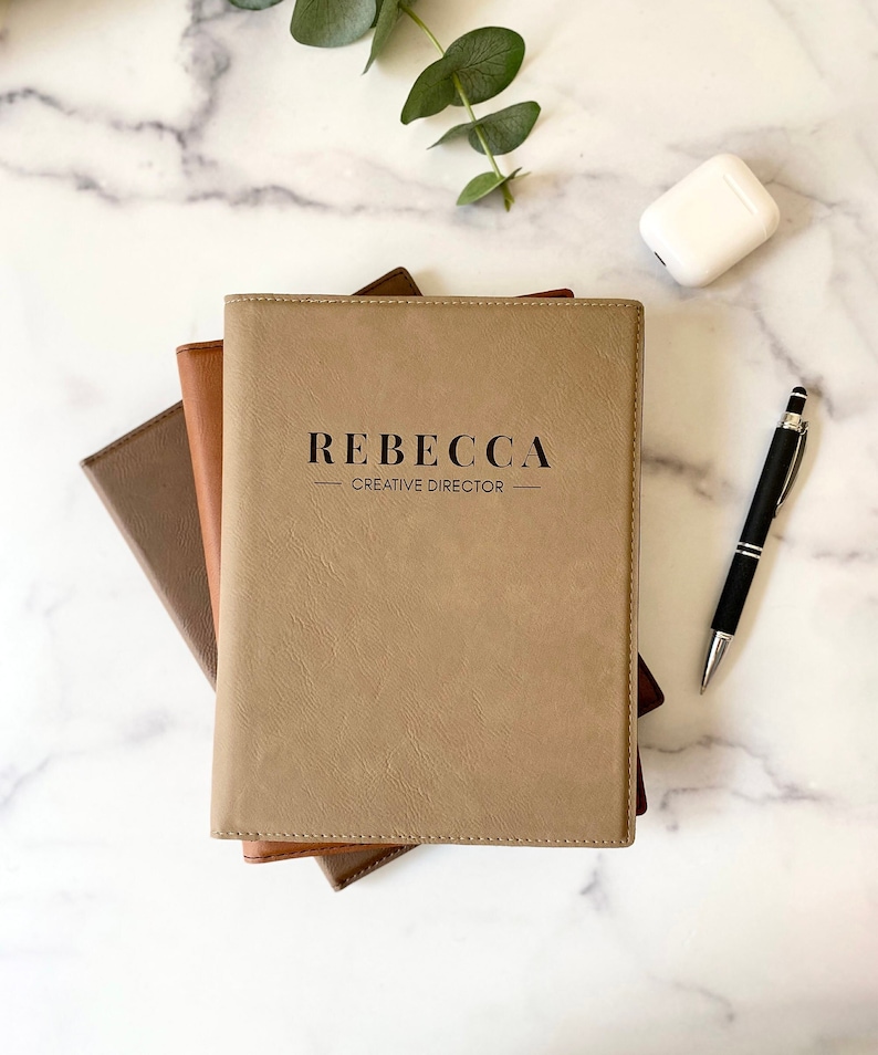 Peut inclure: Une pile de trois journaux reli&eacute;s en cuir marron avec le nom "Rebecca - Directeur de la cr&eacute;ation" grav&eacute; sur le journal du dessus.