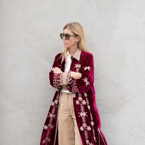 Könnte beinhalten: Burgunderroter Samtmantel mit floraler Stickerei und cremefarbenem Kragen. Getragen mit einem weißen Hemd und beigefarbenen Hosen. Das Outfit wird durch Sonnenbrille und weiße Turnschuhe ergänzt.