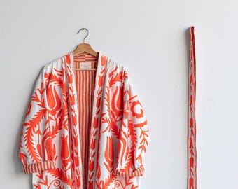 May include: A white robe with a vibrant orange floral pattern, hanging on a wooden hanger. The robe has long sleeves and a matching belt. The interior lining and trim feature a striped design. The robe is approximately 55 inches long.