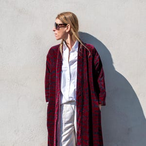 Könnte beinhalten: Ein langer, bordeauxfarbener Samtmantel mit dunklem Muster wird über einem weißen Hemd und einer Hose getragen. Der Mantel reicht bis zu den Knöcheln und hat eine lockere Passform. Das Outfit wird durch weiße Turnschuhe ergänzt.