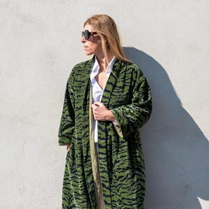 Puede incluir: Una bata larga verde con un estampado de rayas negras de tigre. La bata está abierta, revelando una camisa y pantalones blancos. La persona lleva gafas de sol. La bata está hecha de un material suave, similar al terciopelo.