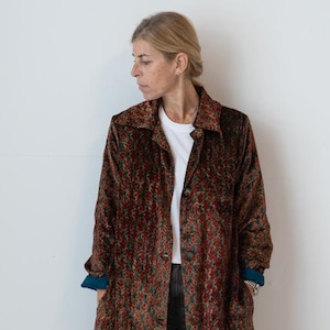 May include: A long, button-down coat with a floral pattern in shades of red, green, and brown. The coat has a collar and long sleeves with blue cuffs. The person is wearing a white shirt and dark trousers. The coat reaches the ankles.