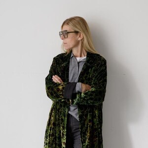 以下が含まれることがあります： 黒と緑のヒョウ柄の長い緑色のベルベットのローブ。ローブは灰色のシャツと濃い色のジーンズの上に着用されています。その人は白いスニーカーを履いています。
