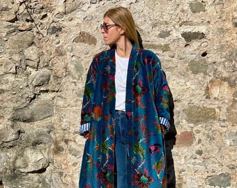 May include: A long teal blue velvet robe with a floral print. The robe has a shawl collar and long sleeves with white and blue striped cuffs. The robe is worn over a white t-shirt and blue jeans.