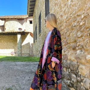 May include: A long black velvet robe with a colorful floral pattern. The robe has a wide collar and long sleeves. The person wearing the robe is standing in front of a stone wall.