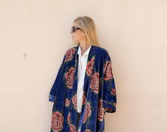 May include: A long blue velvet robe with a floral print of pink roses. The robe has a shawl collar and long sleeves. The model is wearing the robe with a white shirt and grey pants.