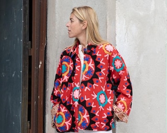 May include: A woman wearing a red, black, and blue patterned jacket with white pants. The jacket has a floral design with a black background.