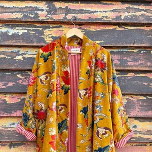 May include: A mustard yellow velvet kimono with a floral print and red and white striped lining. The kimono is hanging on a wooden hanger against a weathered wooden wall.