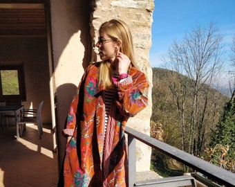 Puede incluir: Una mujer lleva un kimono largo de color naranja con estampado floral y una camisa a rayas rojas y blancas debajo. Está de pie en un balcón con vistas a un bosque y montañas en el fondo.