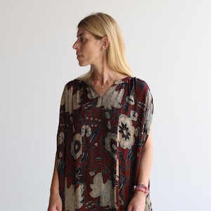 May include: A burgundy, knee-length dress with a floral pattern in beige, navy, and white. The dress has a V-neck and short sleeves. The model is wearing a bracelet and a necklace.