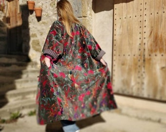 Puede incluir: Una bata larga y fluida con un vibrante estampado floral en tonos rojos, rosas y verdes sobre un fondo oscuro. La bata tiene mangas anchas y un puño a rayas en contraste. La prenda se usa al aire libre.