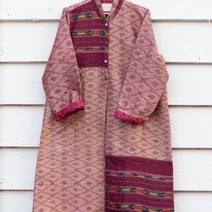 May include: A long, quilted coat with a burgundy and gold patterned design. The coat has a mandarin collar and a button closure. The sleeves are long and have a burgundy and gold patterned trim.