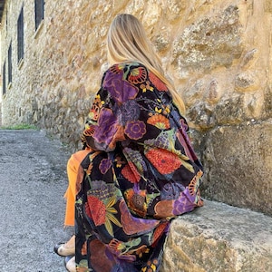 May include: A long black velvet robe with a colorful floral print. The robe has a wide collar and long sleeves. The person wearing the robe is sitting on a stone wall.