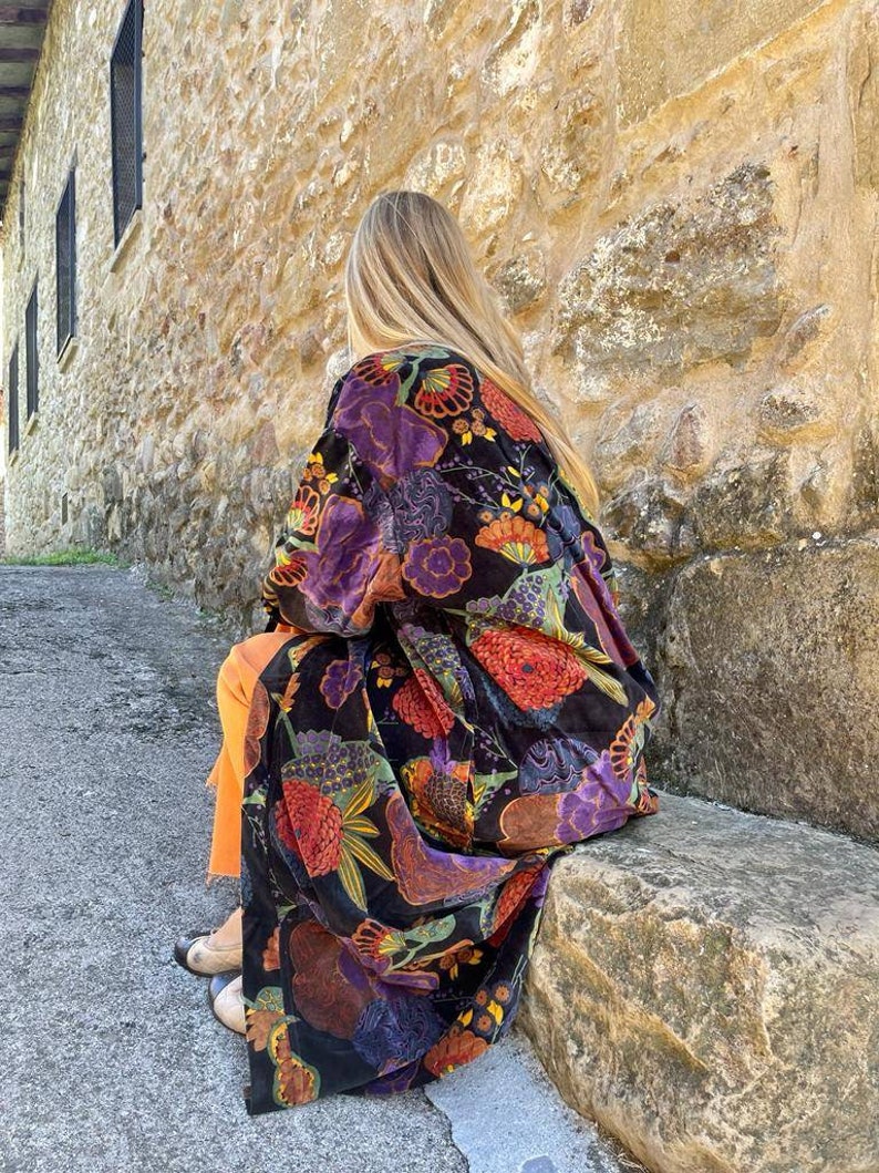 May include: A long black velvet robe with a colorful floral print. The robe has a wide collar and long sleeves. The person wearing the robe is sitting on a stone wall.