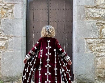 May include: A long, burgundy velvet robe with white floral embroidery. The robe has a long, flowing silhouette and is open in the front. The robe is worn by a person with curly hair, but the person's face is not visible.
