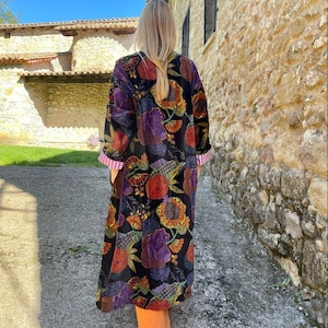 May include: A long black velvet robe with a colorful floral print. The robe has long sleeves and a shawl collar. The robe is worn by a person standing in front of a stone wall.