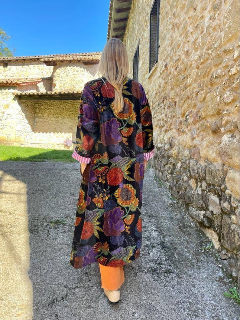 May include: A long black velvet robe with a colorful floral print. The robe has long sleeves and a shawl collar. The robe is worn by a person standing in front of a stone wall.