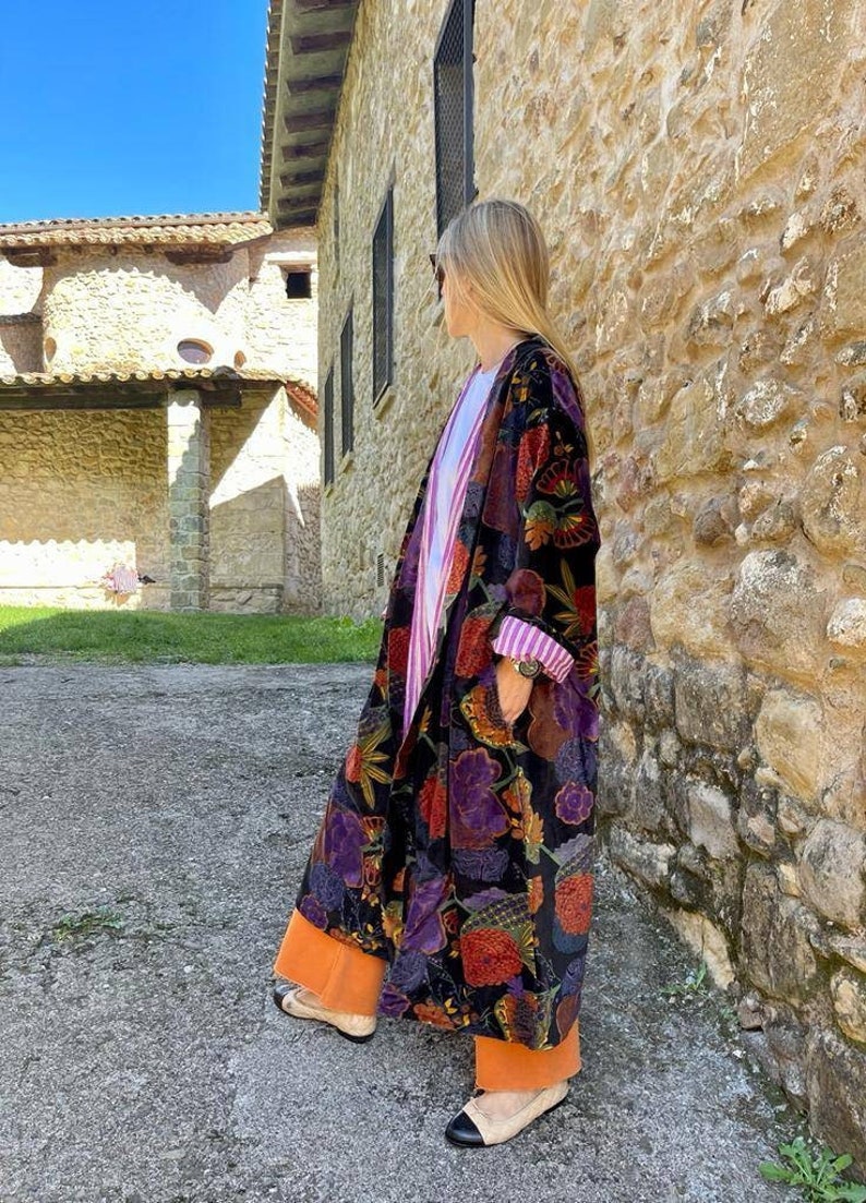 May include: A long black velvet robe with a colorful floral pattern. The robe has a wide collar and long sleeves. The person wearing the robe is standing in front of a stone wall.