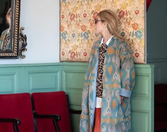 May include: A woman wearing a blue and orange patterned coat stands in front of a floral wallpaper. She is wearing a leopard print sweater and a white shirt. The coat has a long, flowing design and is made of a soft, luxurious fabric.