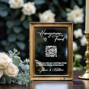 May include: A gold-framed sign with the words "Honeymoon Fund" and a QR code. The sign is on a dark wooden surface. A lit candle in a gold candlestick and a bouquet of white roses are also visible in the image.