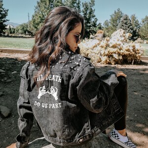 K&ouml;nnte beinhalten: Eine schwarze Jeansjacke mit Perlenbesatz und einem wei&szlig;en Aufdruck auf der R&uuml;ckseite, der "Till Death Do Us Part" lautet, mit zwei Skeletth&auml;nden, die sich am kleinen Finger halten.