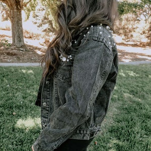 K&ouml;nnte beinhalten: Eine schwarze Jeansjacke mit Perlenbesatz an den Schultern. Die Jacke hat einen klassischen Jeansjackenstil mit Knopfleiste und zwei Taschen.