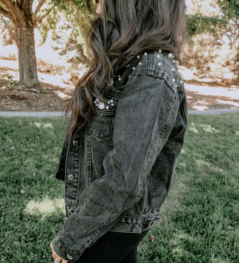 K&ouml;nnte beinhalten: Eine schwarze Jeansjacke mit Perlenbesatz an den Schultern. Die Jacke hat einen klassischen Jeansjackenstil mit Knopfleiste und zwei Taschen.