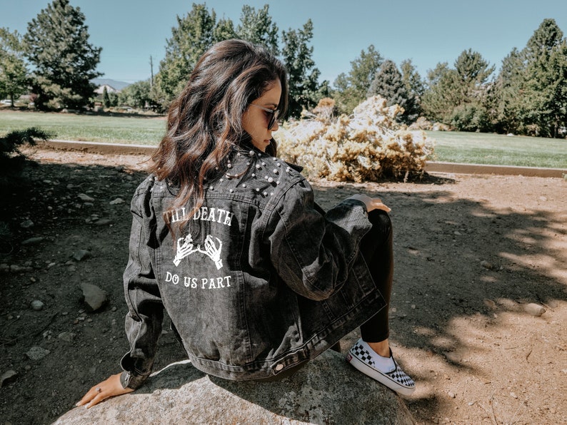 K&ouml;nnte beinhalten: Eine schwarze Jeansjacke mit Perlenbesatz und einem wei&szlig;en Aufdruck auf der R&uuml;ckseite, der "Till Death Do Us Part" lautet, mit zwei Skeletth&auml;nden, die sich am kleinen Finger halten.