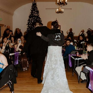 K&ouml;nnte beinhalten: Eine Braut und ein Br&auml;utigam gehen beim Empfang ihrer Hochzeit den Gang entlang. Die Braut tr&auml;gt ein wei&szlig;es Spitzenhochzeitskleid und eine Jeansjacke mit dem Text "Love Us Pet" auf dem R&uuml;cken. Der Br&auml;utigam tr&auml;gt einen schwarzen Anzug. Die G&auml;ste sitzen an Tischen mit violetten Tischdecken und beobachten das Paar.