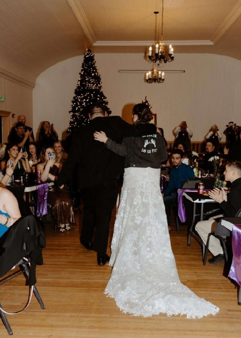 K&ouml;nnte beinhalten: Eine Braut und ein Br&auml;utigam gehen beim Empfang ihrer Hochzeit den Gang entlang. Die Braut tr&auml;gt ein wei&szlig;es Spitzenhochzeitskleid und eine Jeansjacke mit dem Text "Love Us Pet" auf dem R&uuml;cken. Der Br&auml;utigam tr&auml;gt einen schwarzen Anzug. Die G&auml;ste sitzen an Tischen mit violetten Tischdecken und beobachten das Paar.