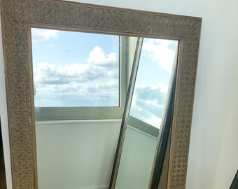 May include: A large, rectangular mirror with a gold-toned frame. The frame has a woven pattern and a slightly distressed finish. The mirror is reflecting a view of a cloudy sky and a window.