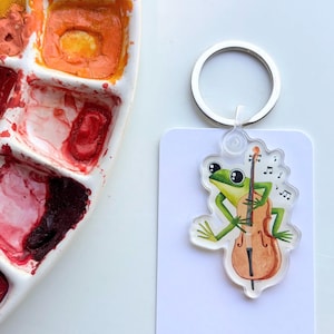 May include: A clear acrylic keychain featuring a green frog playing a brown cello with musical notes floating above. The keychain is attached to a silver metal ring and mounted on a white rectangular card labeled Owlfeathers Watercolor.