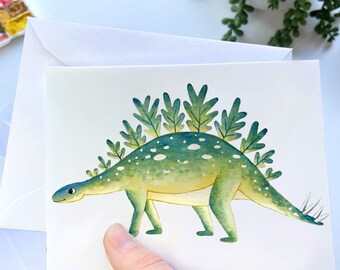 May include: A white greeting card featuring a watercolor illustration of a green dinosaur with leaf-shaped plates along its back. The dinosaur has a yellow underbelly, white spots, and small spikes on its tail. The card is blank inside.