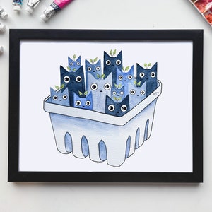May include: Watercolor painting of a collection of blue cats in a white carton. The painting is framed in black and displayed on a white surface.  Various paint tubes and a watercolor paint palette are also visible. The overall theme is whimsical and cute.