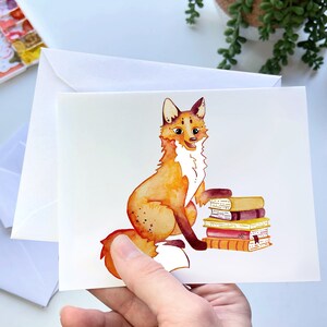 May include: A watercolor illustration of a fox sitting with a stack of books. The fox is orange and white with a fluffy tail. The books are red, orange, and yellow.