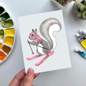 May include: A watercolor illustration of a gray squirrel skiing downhill, wearing pink goggles and a scarf. The squirrel is on pink skis and holding ski poles. The artwork is on a white card, with paint tubes and a palette in the background.