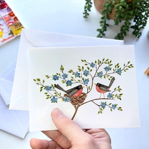 May include: A watercolor painting of two robins on a branch with blue flowers. One robin is sitting in a nest and the other is perched on a branch.
