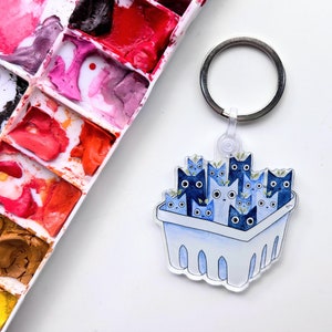 May include: A blue and white key chain with a basket full of blue cats. The key chain has a silver ring.