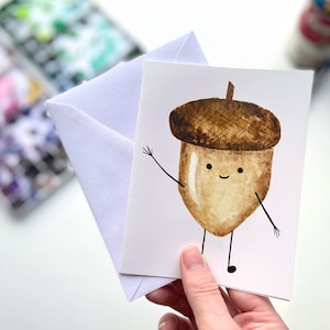 May include: A white card with a watercolor illustration of a smiling acorn with brown and tan colors. The acorn has two small black legs and is waving with one arm.