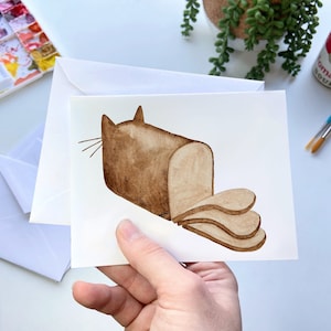 May include: A watercolor illustration of a brown cat shaped like a loaf of bread with slices cut out of the side. The cat is on a white card.