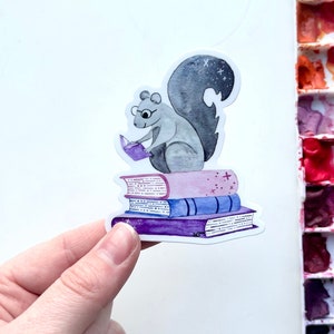 May include: A sticker featuring a gray squirrel wearing glasses and reading a purple book while sitting atop a stack of books. The books are purple, blue, and pink with star accents. The squirrel has a fluffy tail with a starry design.