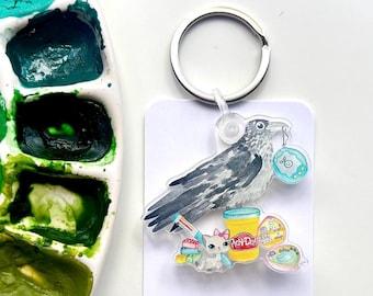 May include: A clear acrylic keychain featuring a watercolor-style illustration of a crow holding a blue toy. The keychain also includes images of Play-Doh, a cat, and other toys. A silver key ring is attached to the top.