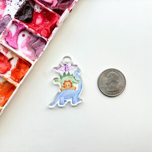 May include: A clear acrylic charm with a watercolor illustration of four dinosaurs stacked on top of each other. The dinosaurs are blue, green, orange, and purple. The charm is about the size of a quarter.