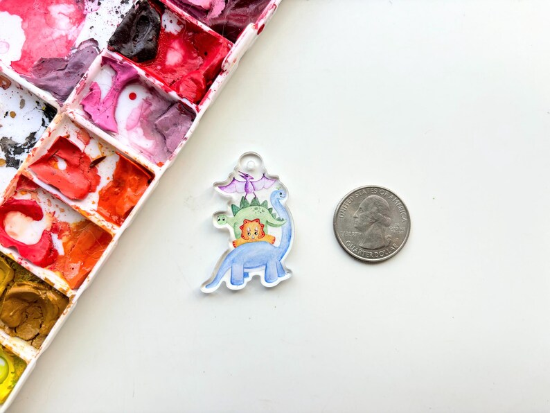 May include: A clear acrylic charm with a watercolor illustration of four dinosaurs stacked on top of each other. The dinosaurs are blue, green, orange, and purple. The charm is about the size of a quarter.