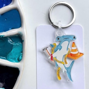 May include: A novelty keychain featuring a cartoon hammerhead shark with a construction theme. The shark is blue and white, holding a hammer and wearing a traffic cone. Yellow caution tape wraps around the shark. A silver keyring is attached.