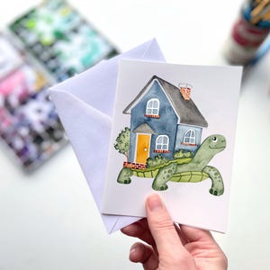 May include: A watercolor greeting card featuring a blue house with a gray roof and orange door, carried by a green turtle. The card is held with a white envelope. Watercolor paints and brushes are in the background.