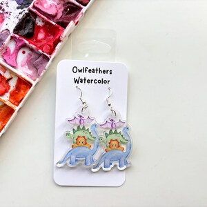 May include: A pair of dangle earrings featuring a watercolor illustration of dinosaurs. The earrings are made of clear acrylic and have silver hooks. The illustration features a blue dinosaur with a green dinosaur and a yellow dinosaur on top. The text "Owlfeathers Watercolor" is printed on the card.