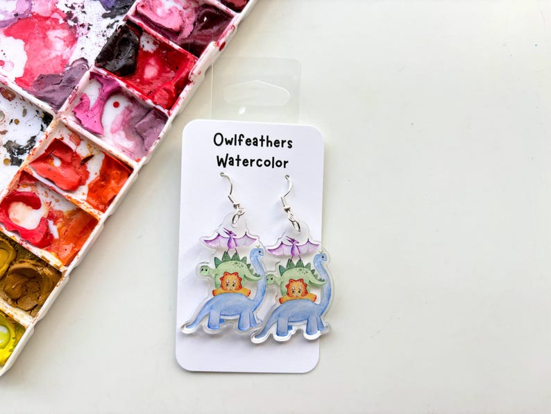 May include: A pair of dangle earrings featuring a watercolor illustration of dinosaurs. The earrings are made of clear acrylic and have silver hooks. The illustration features a blue dinosaur with a green dinosaur and a yellow dinosaur on top. The text "Owlfeathers Watercolor" is printed on the card.