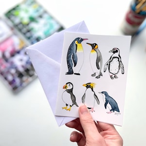 May include: A watercolor illustration of six different penguin species, including a puffin, on a white card. The penguins are depicted in various poses, with some standing upright and others swimming. The illustration is done in a realistic style, with detailed features and vibrant colors.